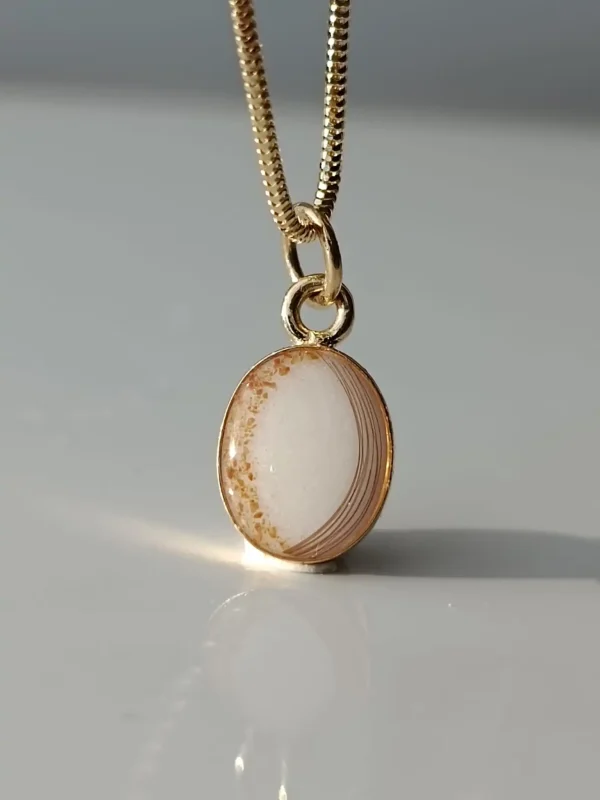 A gold pendant necklace featuring an oval polished stone with natural brown and white layered patterns is displayed on a smooth, reflective surface. The stone is framed in gold, enhancing its elegant appearance.