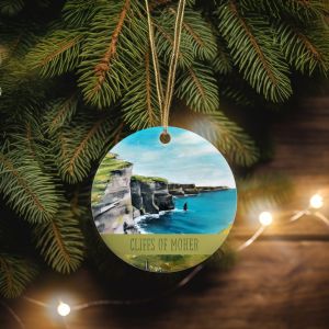 A close-up of a Christmas tree with a round ornament depicting the Cliffs of Moher. The ornament features a painted seascape with cliffs and ocean, hanging among lush green pine needles, with soft fairy lights in the background.