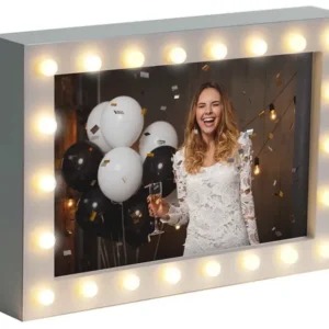 A woman in a white dress smiles while holding a champagne glass, surrounded by black and white balloons, with confetti in the air. The scene is framed by a lighted marquee-style frame with round bulbs.