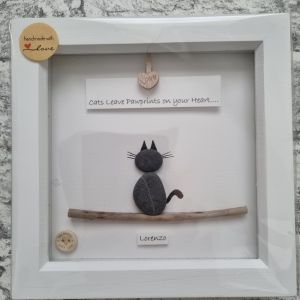 A framed artwork featuring a black cat figurine made from stones and wood. Above the cat is the text "Cats Leave Pawprints on your Heart..." and below, the name "Lorenzo." A small tag reads "handmade with love.