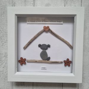 Framed artwork with driftwood and pebbles forming a simple house shape, featuring a small pebble dog inside. A heart with "Love" sits at the peak and the text reads, "A House is not a Home Without Dogs!.