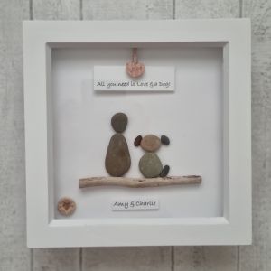 A framed art piece featuring pebble figures of a person and a dog sitting on a twig. Above, a sign reads, "All you need is Love & a Dog!" A small heart hangs at the top, and "Amy & Charlie" is written at the bottom.