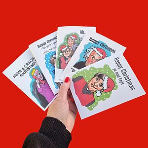 A hand holding several humorous Christmas cards against a red background. The cards feature festive illustrations and playful messages like "Happy Christmas ya mate!" and "Have a crackin' Christmas!.