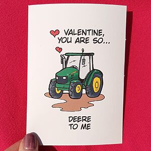 A red background with a greeting card featuring a green tractor illustration. Above the tractor, the text reads "Valentine, you are so..." with hearts, and below it says "Deere to me." A hand is holding the card.