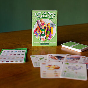 A card game titled "Exploring Ireland" is displayed on a wooden table. The game box shows illustrations of people and landmarks. Various playing cards, featuring images and text, are spread out around the box.