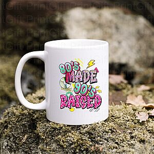 A white mug on a stone surface with moss and fallen leaves, featuring colorful graffiti-style text saying "80's Made 90's Raised" with retro graphics.