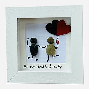 A framed artwork featuring two pebble figures holding hands. The figure on the left holds three heart-shaped balloons. Below them, text reads "All you need is love..." with two heart symbols. The frame is white.